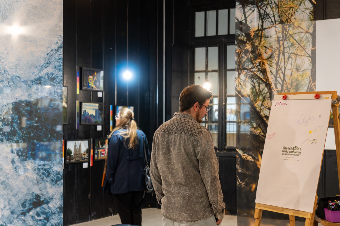 Visitantes observam as fotografias da exposição Cidade, Cor e Ambiente na Galeria Estação da Arte, em Passo Fundo, ao lado de um painel interativo.